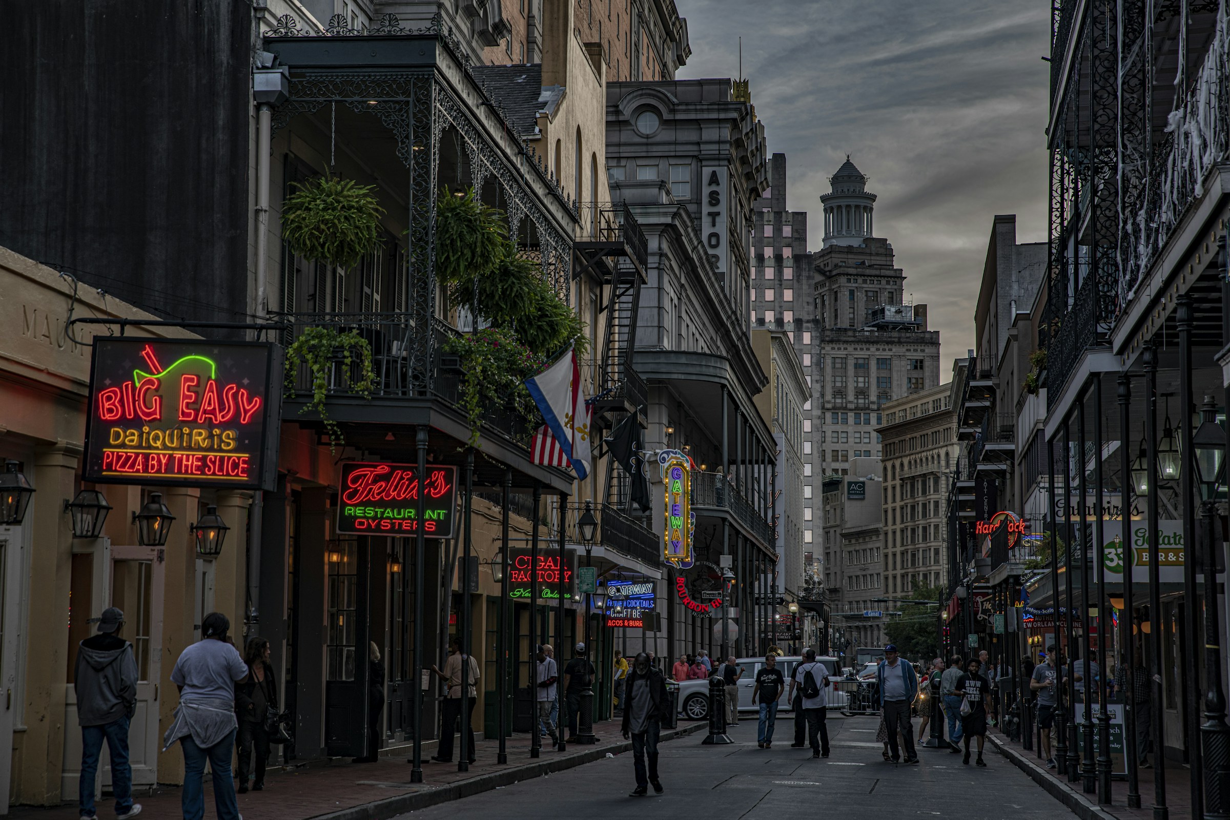 Pedestrians walking down Bourbon Street in the French Quarter at dusk, with the New Orleans CBD skyline in the distance