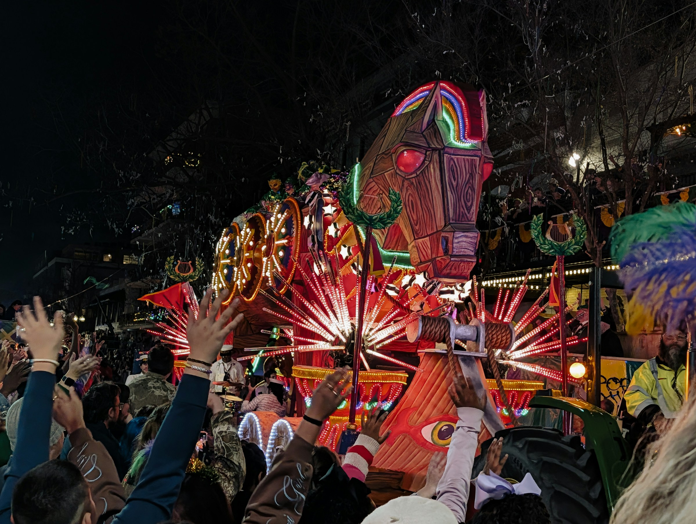 Parade-goers with arms raised as a lit Mardi Gras float rolls past on the New Orleans parade route